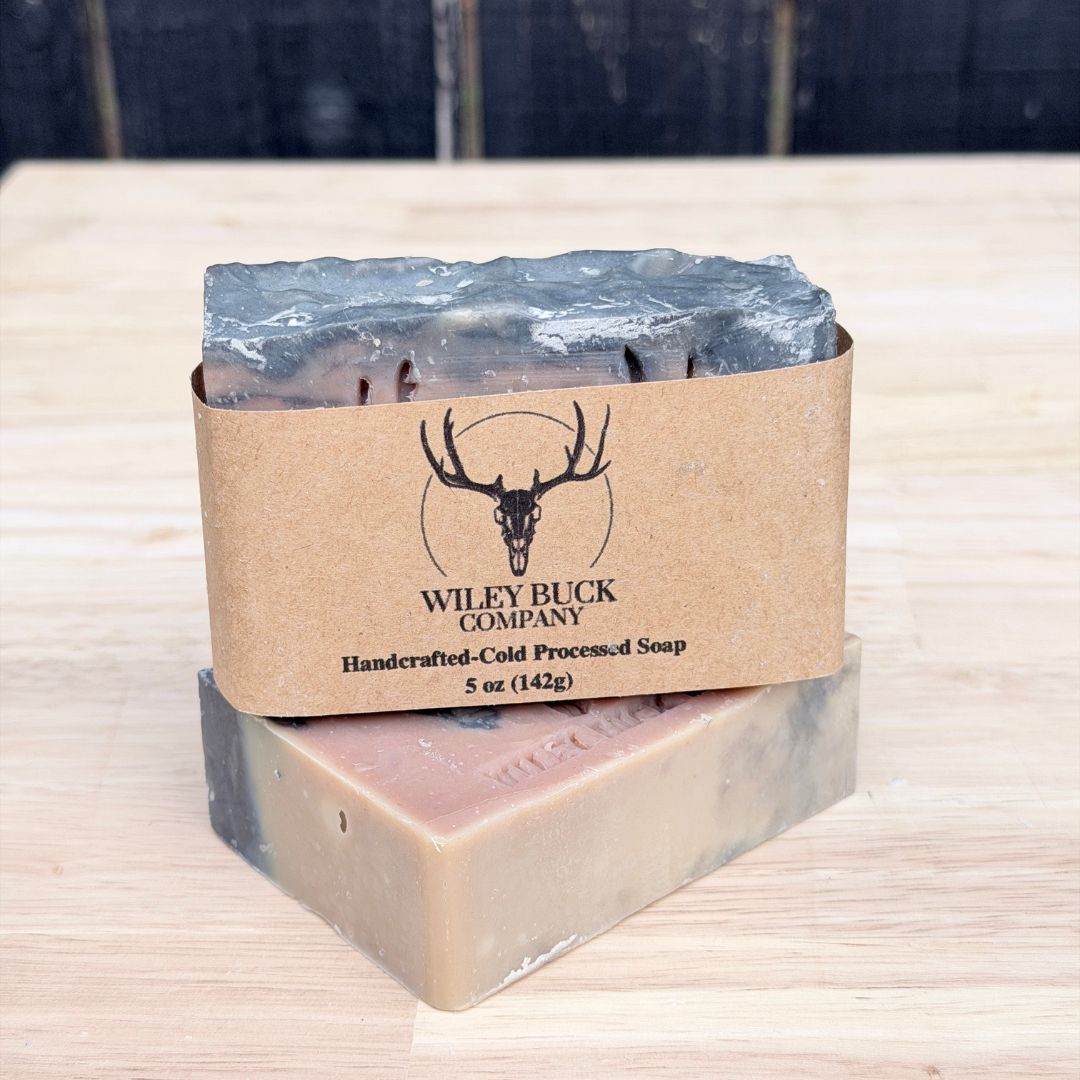 Two bars of soap with a brown paper wrap featuring the Wiley Buck Company logo on a wooden surface.