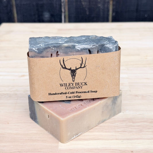 Two bars of soap with a brown paper wrap featuring the Wiley Buck Company logo on a wooden surface.