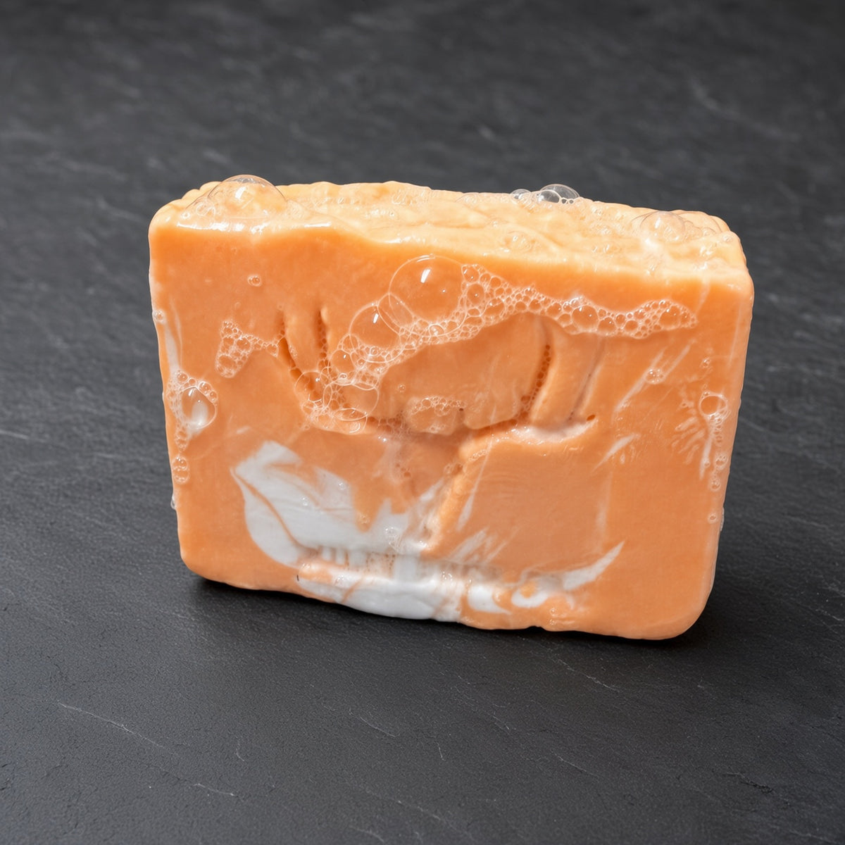A rectangular bar of orange soap with a white marbled swirl pattern and visible soap bubbles on top, resting on a dark gray slate surface.