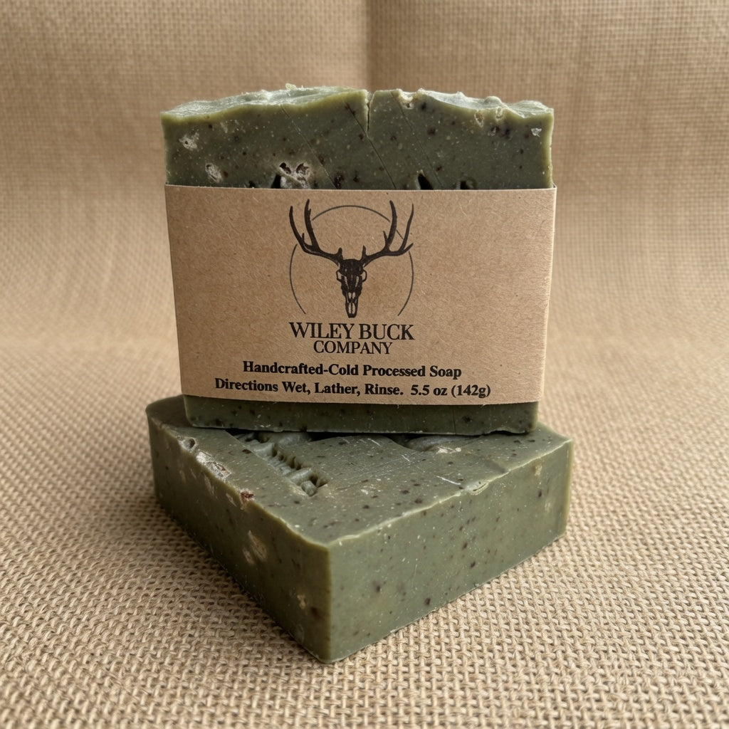 Two bars of handmade soap in a sage green color with visible flecks, stacked on a beige burlap background. The top bar has a kraft paper label wrapped around it with the brand name 'Wiley Buck Company' and a deer skull logo.