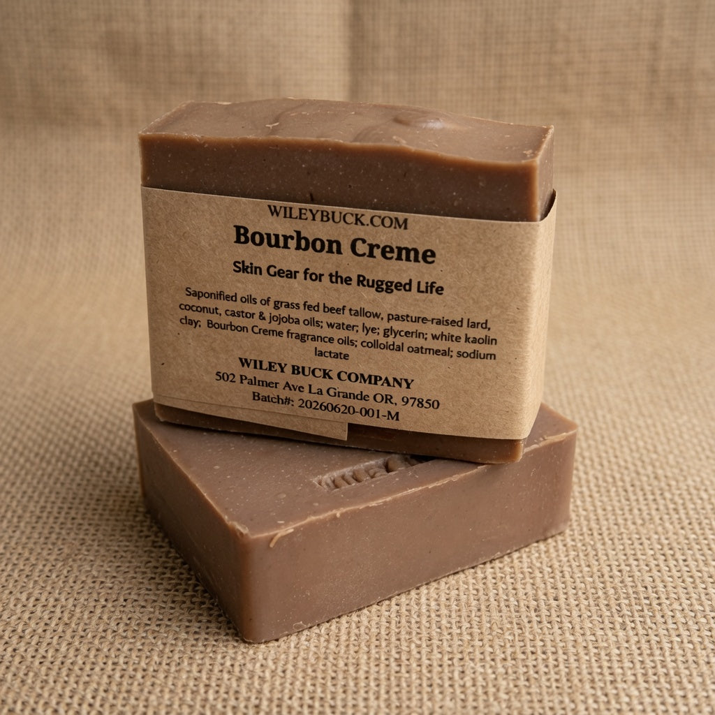 Two rectangular bars of Wiley BUCK Bourbon Creme soap stacked on a textured beige burlap background. The top bar is slightly offset and angled, showing a kraft paper label with black text, while the bottom bar is plain and shows the soap's smooth, matte brown surface.
