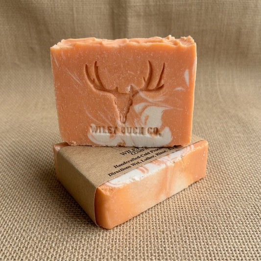 A bar of soap in a orange and white marbled pattern, embossed with a deer head logo and the brand name 'WILEY BUCK CO.', displayed on top of its kraft paper packaging against a neutral burlap background.