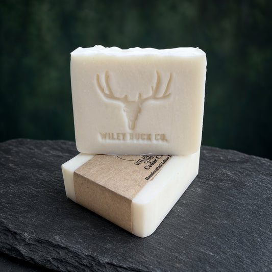 Two rectangular bars of off-white soap stacked on a dark slate surface, with the top bar embossed with a deer head logo and the words 'WILEY BUCK CO.' The lower bar is wrapped in a kraft paper band.
