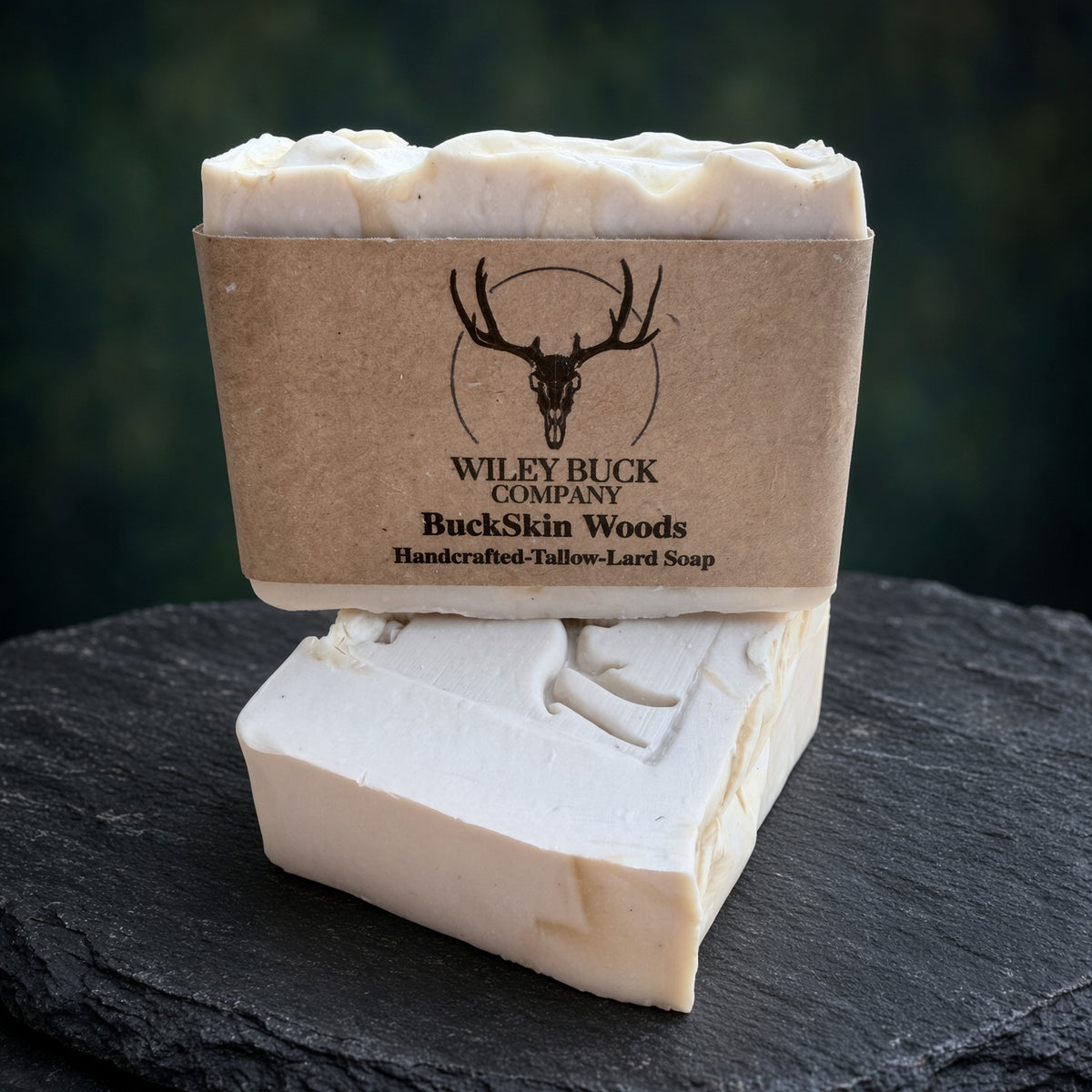 A handmade bar of soap shown unwrapped from a kraft paper sleeve, with the branded sleeve standing behind it on a dark slate surface. The soap is a warm off-white/cream color with a carved deer design on the front.