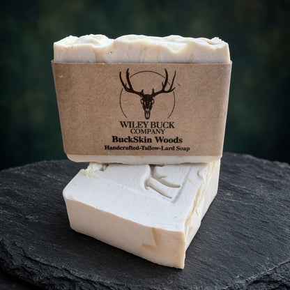 A handmade bar of soap shown unwrapped from a kraft paper sleeve, with the branded sleeve standing behind it on a dark slate surface. The soap is a warm off-white/cream color with a carved deer design on the front.