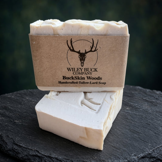 A handmade bar of soap shown unwrapped from a kraft paper sleeve, with the branded sleeve standing behind it on a dark slate surface. The soap is a warm off-white/cream color with a carved deer design on the front.