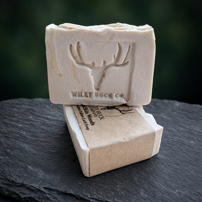 A rectangular bar of natural soap in a light cream color, embossed with a deer head antler logo and the brand name 'WILEY BUCK CO.' resting on top of its kraft paper packaging, set on a dark slate surface with a blurred dark green background.