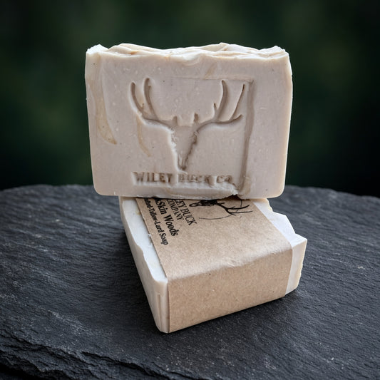 A rectangular bar of natural soap in a light cream color, embossed with a deer head antler logo and the brand name 'WILEY BUCK CO.' resting on top of its kraft paper packaging, set on a dark slate surface with a blurred dark green background.