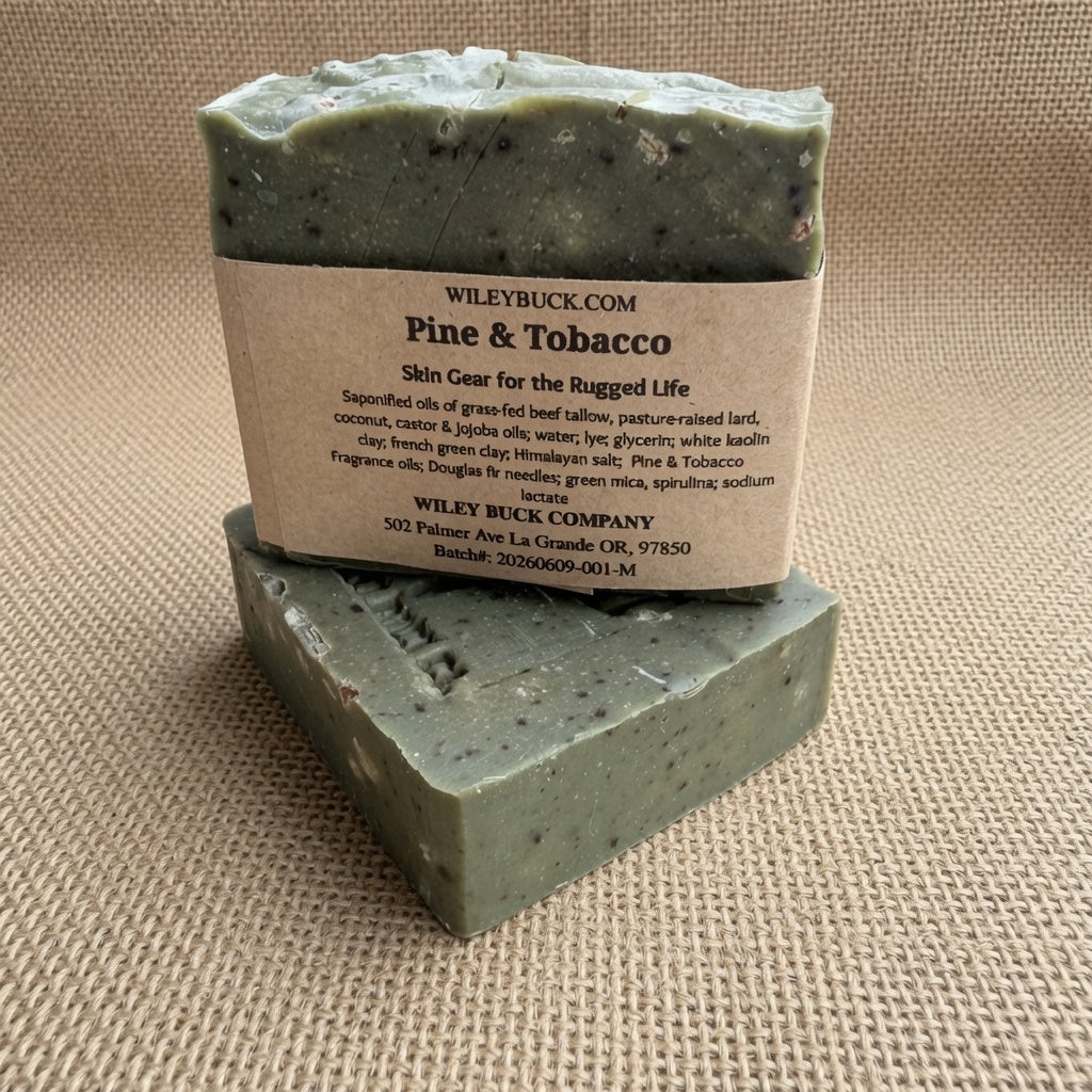 Two rectangular bars of WileyBuck.com Pine & Tobacco skin gear soap stacked on a textured beige fabric background.