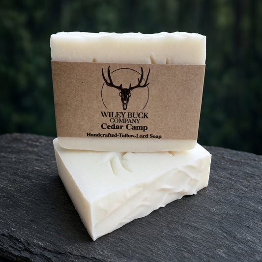 Two bars of Wiley Buck Company Cedar Camp handcrafted tallow-and-lard soap, one wrapped in a brown kraft paper label with a deer skull logo, displayed on a dark slate surface with a blurred forest background.