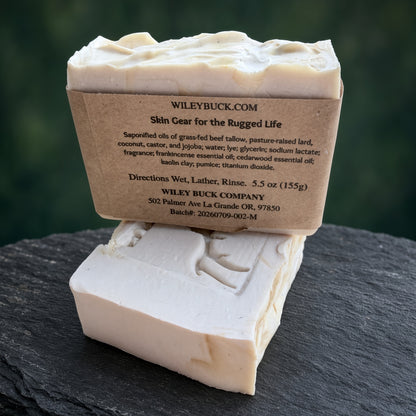 Two rectangular bars of Wiley Buck Soap Company Skin Gear soap, one standing upright with a kraft paper label and one lying flat, set on a dark slate surface with a blurred green background.