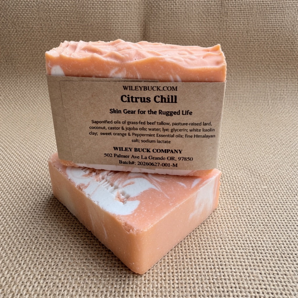 Two rectangular bars of Wiley Buck Company Citrus Chill soap stacked on a textured beige burlap background. The soap has a orange color with white marbling, and is wrapped in a brown kraft paper label.