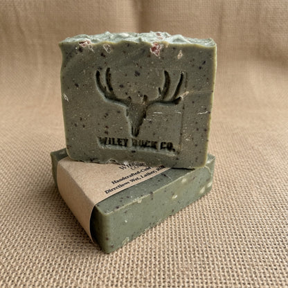 A handmade soap bar in a sage green speckled finish with an embossed deer head logo and the brand name 'Wiley Buck Co.' stacked on top of its kraft paper packaging, set against a neutral burlap background.
