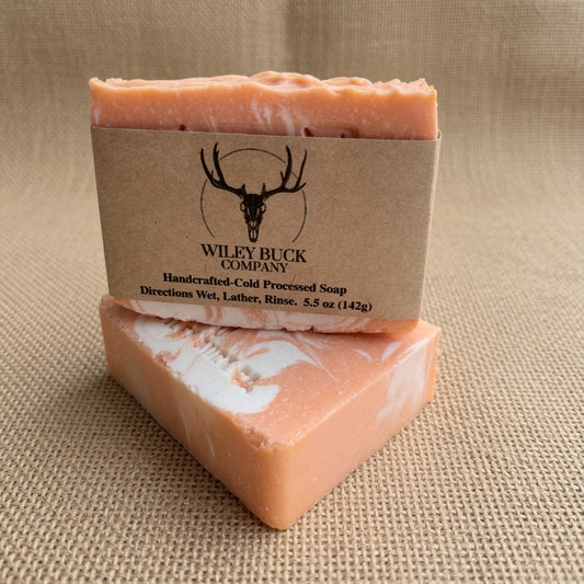 Two bars of Wiley Buck Company handmade cold processed soap, one wrapped in a kraft paper band with a deer skull logo, displayed on a textured beige fabric background.