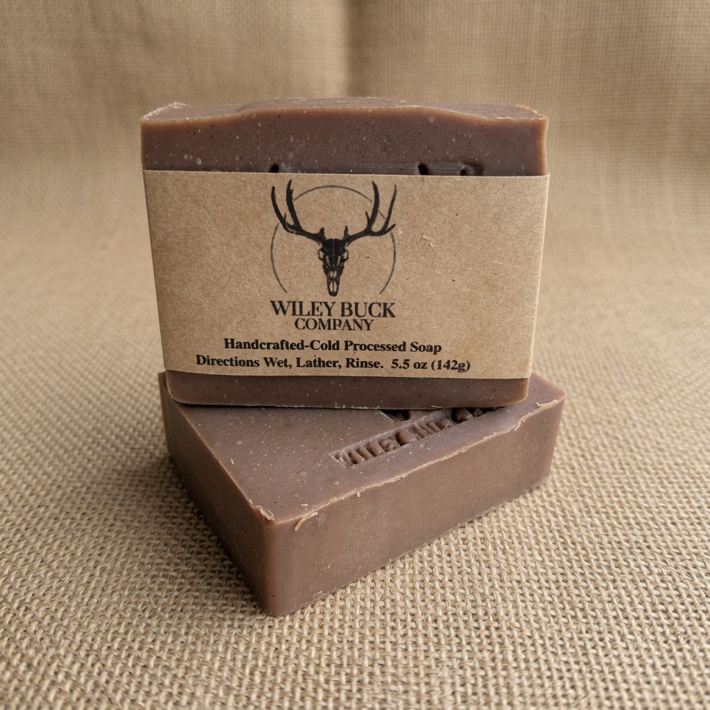Two rectangular bars of Wiley Buck Company handcrafted cold processed soap stacked on a textured beige burlap background. The top bar is wrapped in a kraft paper label featuring a black deer skull logo and brand name.