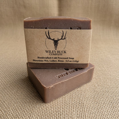 Two rectangular bars of Wiley Buck Company handcrafted cold processed soap stacked on a textured beige burlap background. The top bar is wrapped in a kraft paper label featuring a black deer skull logo and brand name.